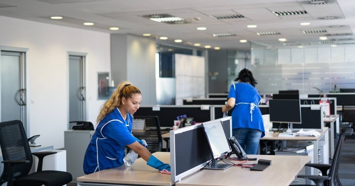 Two people in blue clothing are cleaning computers. There are several desks with computers on them.